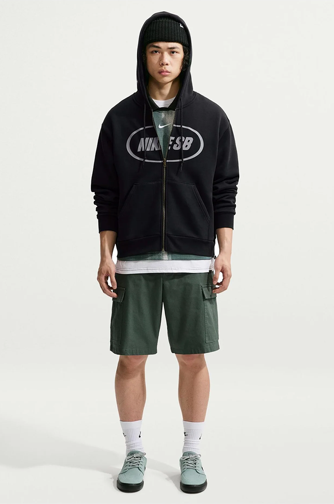 A model wearing a black zip-up hoodie with a white Nike logo, green cargo shorts, a gray t-shirt, white socks, and green sneakers, standing against a light gray studio background.