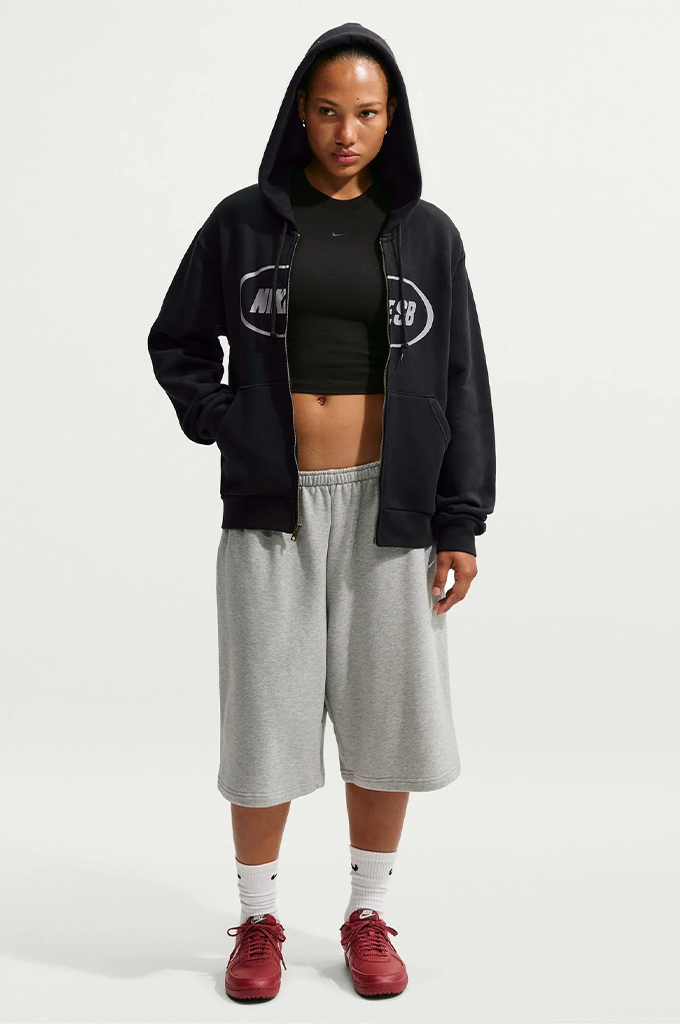 A model stands against a light gray studio background wearing a black Nike zip-up hoodie over a black crop top, paired with light gray sweat shorts, white socks, and red sneakers.