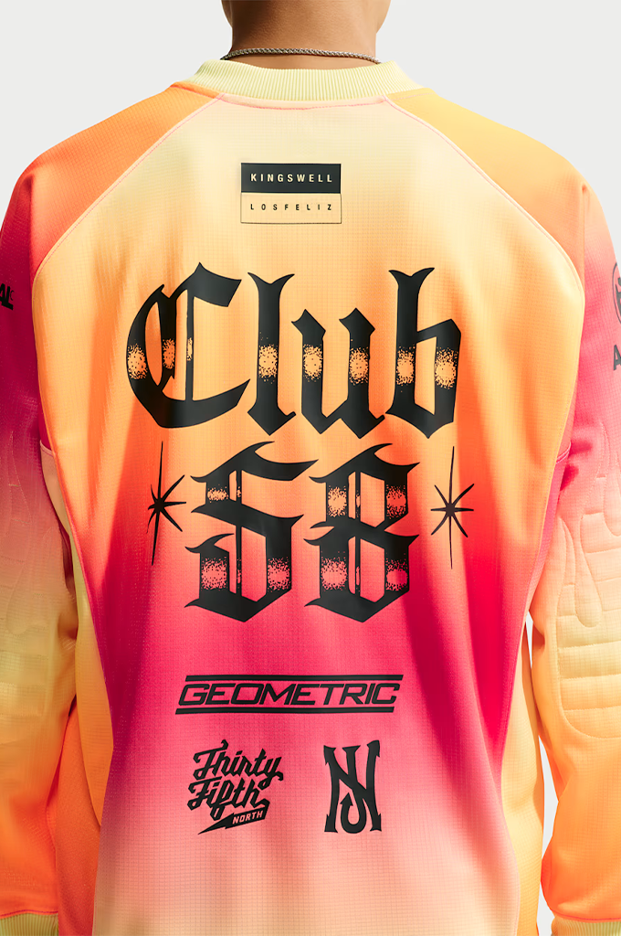 A young man stands centered against a plain light gray background, wearing a vibrant, gradient-colored long-sleeve jersey with a yellow, orange, and hot pink ombré pattern. He wears dark, futuristic wrap-around sunglasses and a thin silver chain necklace.The jersey features a black Swoosh logo on the right chest, a small stylized graphic on the left chest, and a large black graphic in the center depicting a sleeping cherub above the bold block text "SB FC", "LA MÁS FINA", and "FTC".