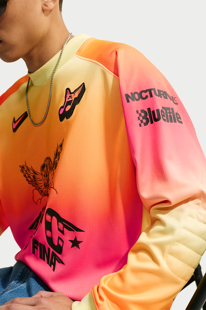 A young man stands centered against a plain light gray background, wearing a vibrant, gradient-colored long-sleeve jersey with a yellow, orange, and hot pink ombré pattern. He wears dark, futuristic wrap-around sunglasses and a thin silver chain necklace. The jersey features a black Swoosh logo on the right chest, a small stylized graphic on the left chest, and a large black graphic in the center depicting a sleeping cherub above the bold block text "SB FC", "LA MÁS FINA", and "FTC".