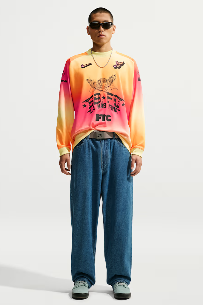 A young man stands centered against a plain light gray background, wearing a vibrant, gradient-colored long-sleeve jersey with a yellow, orange, and hot pink ombré pattern. He wears dark, futuristic wrap-around sunglasses and a thin silver chain necklace. The jersey features a black Swoosh logo on the right chest, a small stylized graphic on the left chest, and a large black graphic in the center depicting a sleeping cherub above the bold block text "SB FC", "LA MÁS FINA", and "FTC".