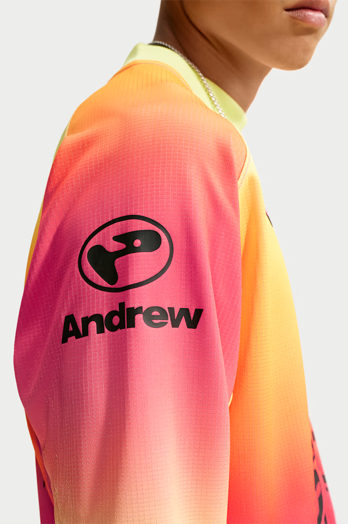 A young man stands centered against a plain light gray background, wearing a vibrant, gradient-colored long-sleeve jersey with a yellow, orange, and hot pink ombré pattern. He wears dark, futuristic wrap-around sunglasses and a thin silver chain necklace. The jersey features a black Swoosh logo on the right chest, a small stylized graphic on the left chest, and a large black graphic in the center depicting a sleeping cherub above the bold block text "SB FC", "LA MÁS FINA", and "FTC".