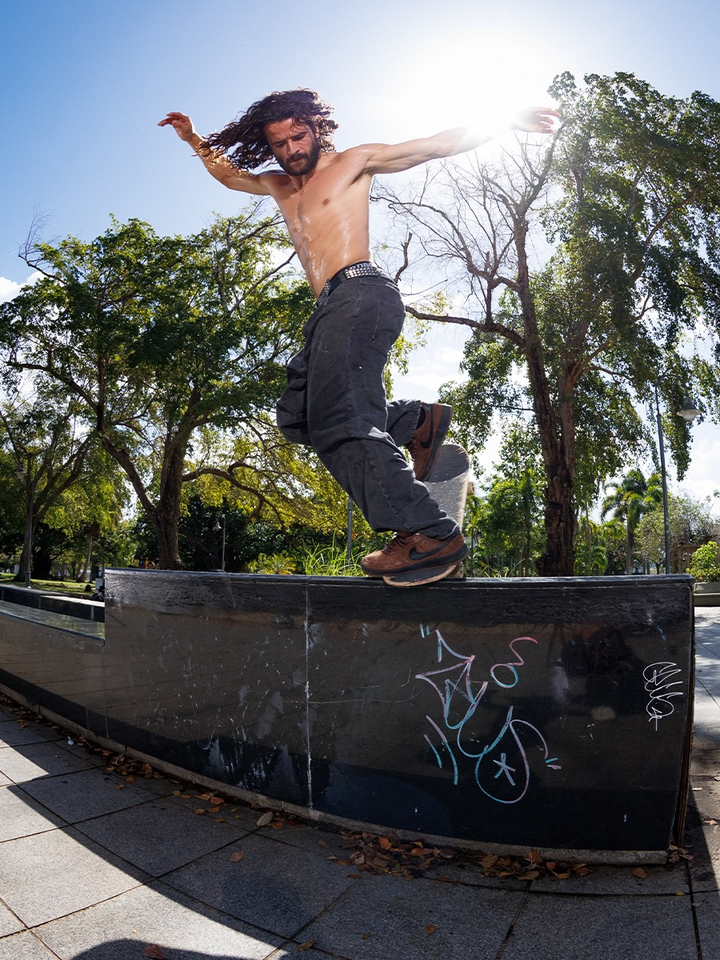 A shirtless man with long dark hair and a beard jumps over a concrete ledge in a park, wearing loose dark pants and brown shoes, with trees and bright sunlight in the background.