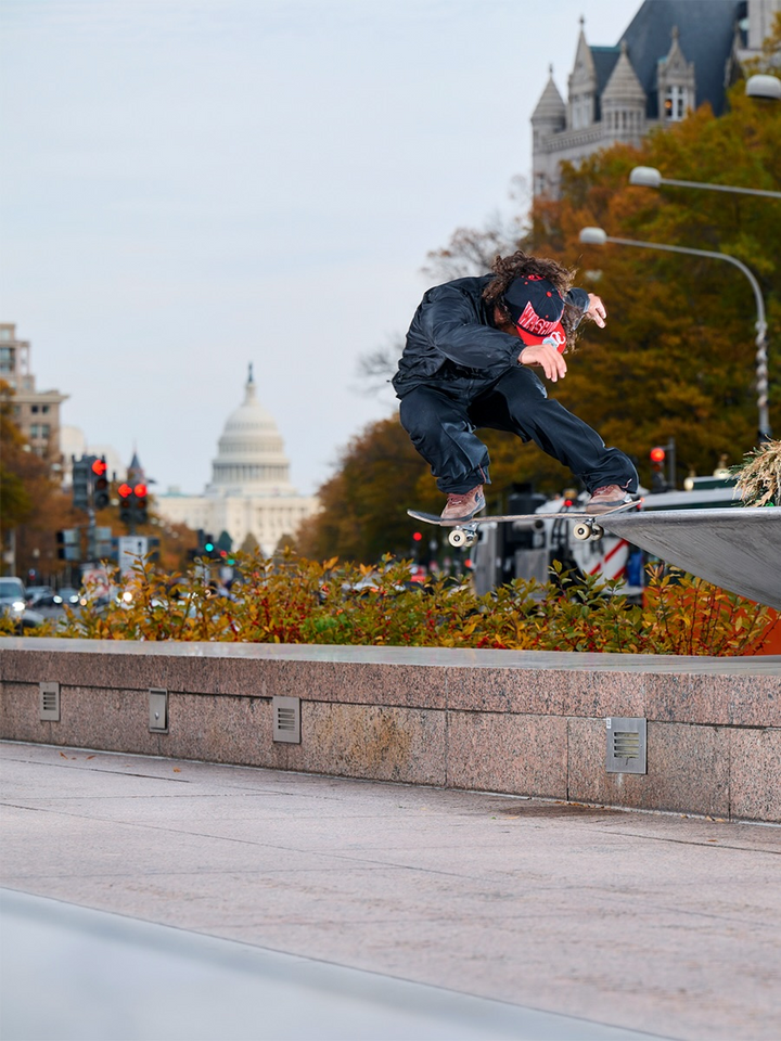 A skateboarder in a black outfit and red bandana jumps a skateboard over a concrete ramp in an urban setting, with the U.S.