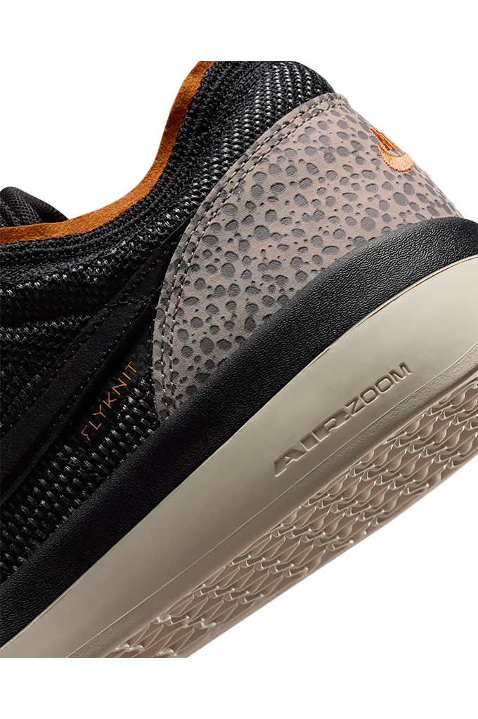 Close-up of a Nike sneaker showing the Flyknit upper, textured gray heel panel, orange heel tab, and beige Air-Zoom sole.#color_cobblestone-black-monarch