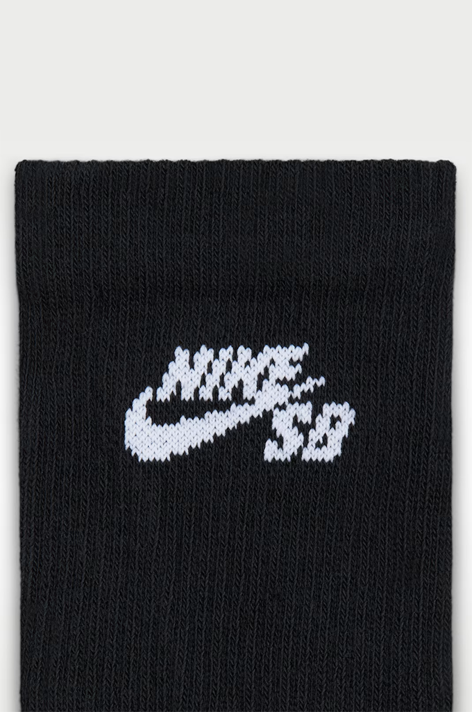 A close-up of a black ribbed knit sock featuring a white knitted Nike SB logo across the front, set against a light gray background.#color_black