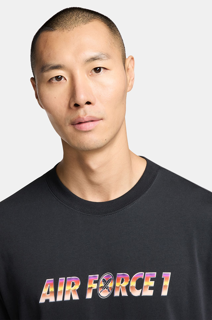 A close-up shot of a man wearing a black crewneck t-shirt with a colorful 'AIR FORCE 1' graphic printed across the chest, set against a plain light gray background.#color_black