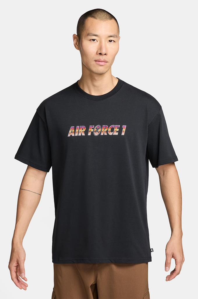 A man wearing a black short-sleeve t-shirt with 'AIR FORCE 1' printed across the chest in a multicolor pattern, paired with brown pants, against a light gray background.#color_black