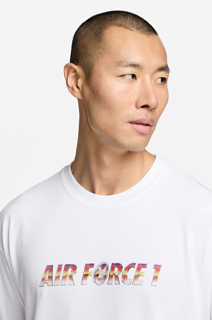 A man wearing a white crewneck t-shirt with 'AIR FORCE 1' printed across the chest in a colorful gradient pattern, set against a plain light gray background.#color_white