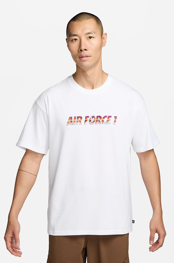 A man wearing a white short-sleeve t-shirt with 'AIR FORCE 1' printed across the chest in a red, orange, and yellow gradient, paired with brown pants, against a light gray background.#color_white