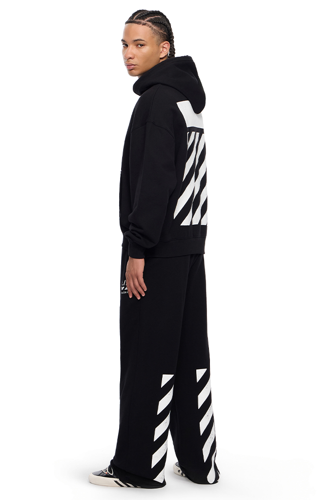 A model wearing a black Off-White tracksuit with signature white diagonal arrow stripes on the back of the hoodie and on the pants, shown from a side profile against a white background.