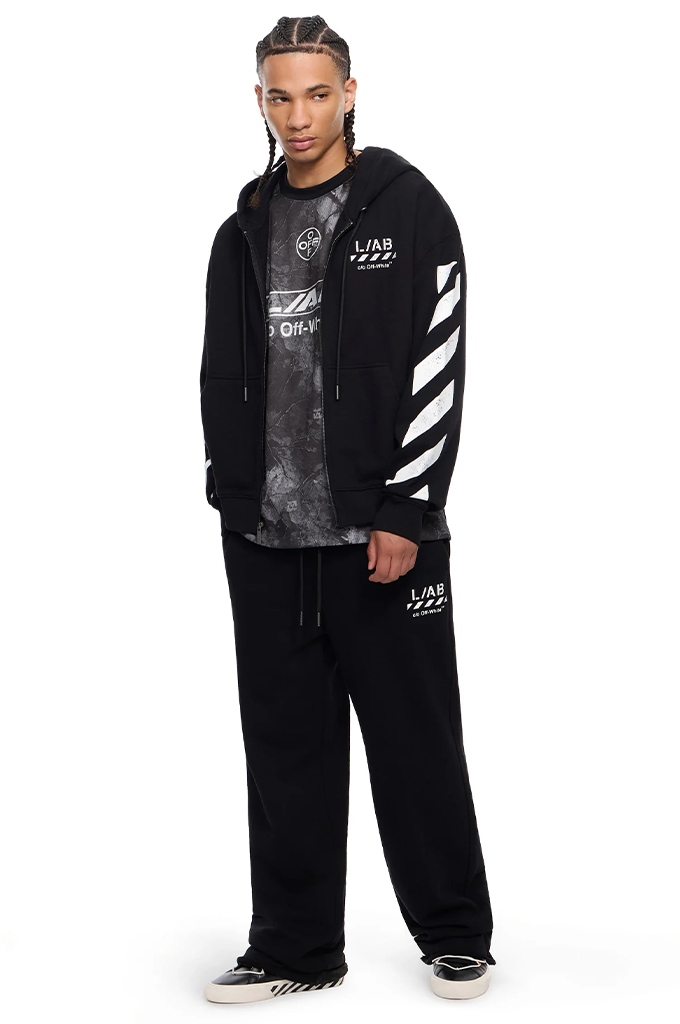 A male model wearing a black streetwear outfit featuring a black hoodie with white diagonal stripes on the sleeve, a black and gray graphic t-shirt, black sweatpants with white graphics, and black and white sneakers, set against a plain white background.