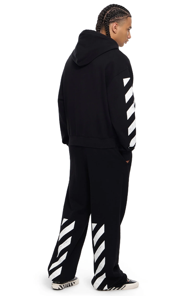 A model shown from the back wearing a black hoodie and matching sweatpants, both featuring the signature white diagonal stripes of Off-White on the sleeves and legs, against a plain white background.