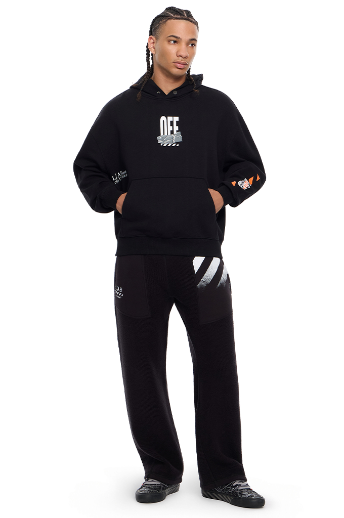 A man modeling a black Off-White streetwear set, featuring a pullover hoodie with a white 'OFF' logo on the chest and matching sweatpants with white diagonal stripes on the thigh.#color_black
