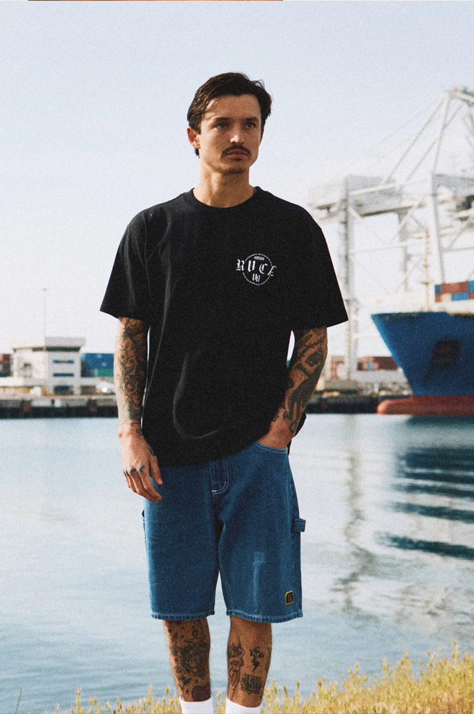A man with tattoos stands by a waterfront wearing a black crewneck t-shirt with a white graphic logo on the chest, paired with blue denim shorts.