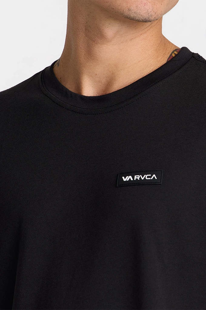 Close-up of a person wearing a black crewneck t-shirt with a small black rectangular patch on the chest featuring the white 'RVCA' logo.