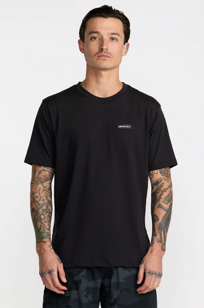 A man wearing a black short-sleeve crewneck t-shirt with a small white Vans logo on the left chest, standing against a light gray studio background.