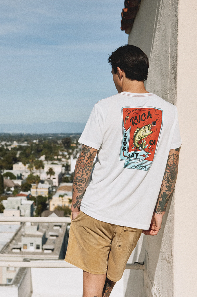 A man wearing a white short-sleeve t-shirt with a large red and yellow graphic print on the back, paired with tan shorts, standing on a balcony with a city view.#color_antique-white