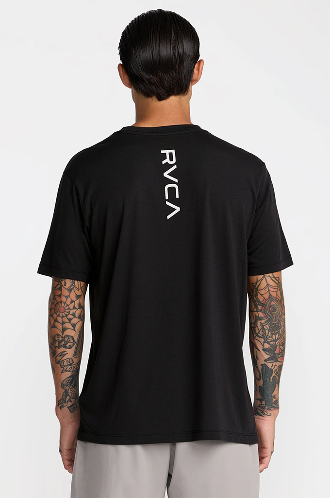 A man viewed from the back wearing a black short-sleeve t-shirt with a vertical white RVCA logo on the upper back, paired with light gray pants, against a plain light gray studio background.