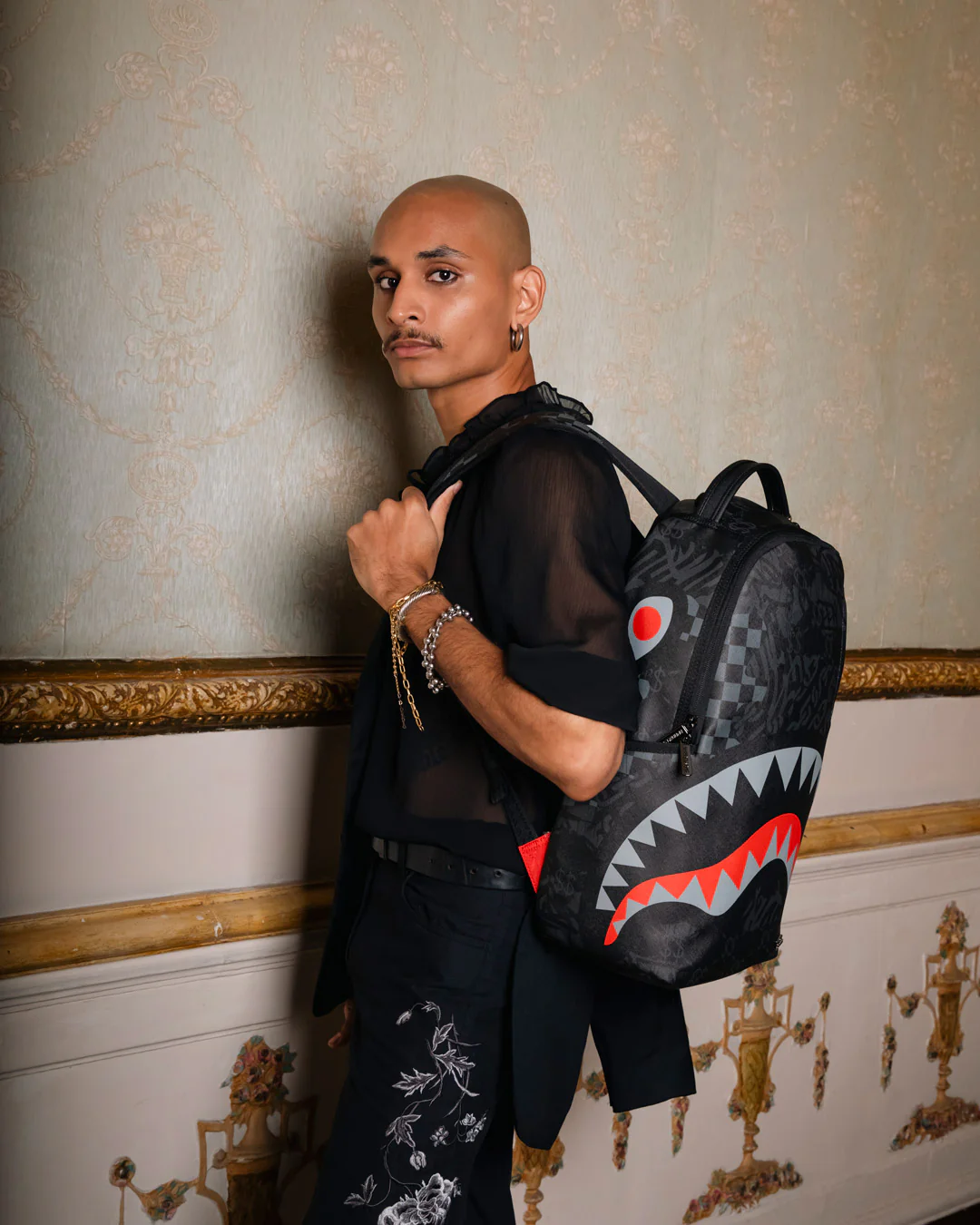 A model wearing a black sheer long-sleeve top and black patterned trousers, carrying a black shark-mouth backpack with red and white details, posed against a classic damask wallpaper wall.