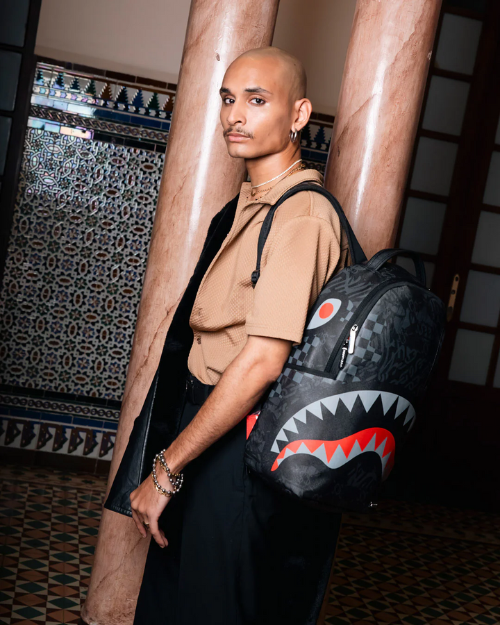 A man wearing a tan short-sleeve shirt and black trousers carries a black shark-face backpack with red, white, and gray details, standing in front of two large marble columns in an indoor setting.