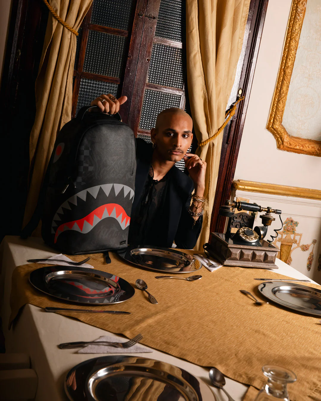 A man in a black shirt sits at a table draped with a tan cloth, holding up a black shark-mouth backpack. The setting is an ornate room with gold curtains, a vintage telephone, and gold-framed wall art.