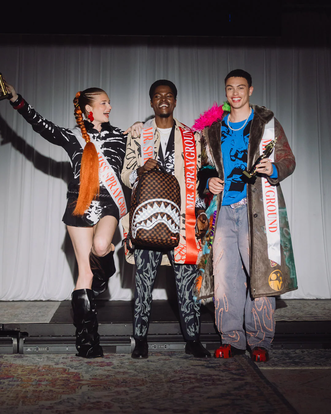 Three models posing together on a stage, each wearing a highly detailed, colorful streetwear outfit with shark-print patterns, sashes, and accessories. The center model holds a brown patterned shoulder bag with a shark mouth design.