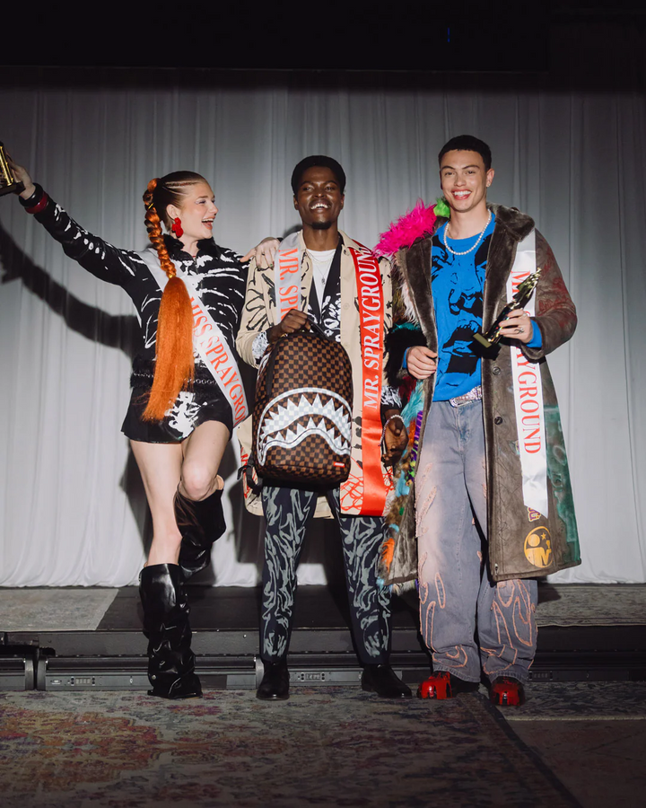 Three models posing together on a stage, each wearing a highly detailed, colorful streetwear outfit with shark-print patterns, sashes, and accessories. The center model holds a brown patterned shoulder bag with a shark mouth design.