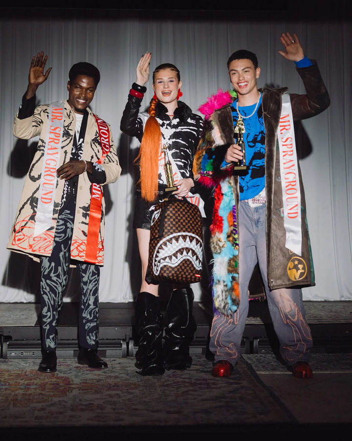 Three individuals on a stage, two with colorful outfits and one holding an award, against a plain background.