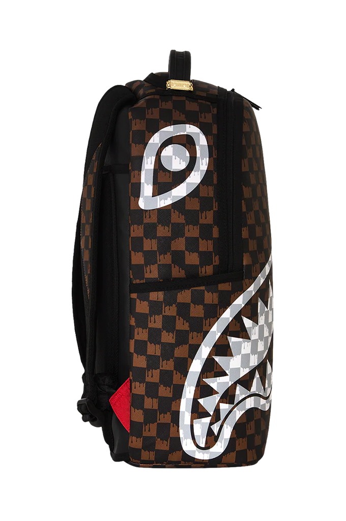 A Sprayground Shark backpack featuring a brown and black checkered pattern, a large white shark mouth graphic across the front, and a small red Sprayground logo tag at the bottom right.