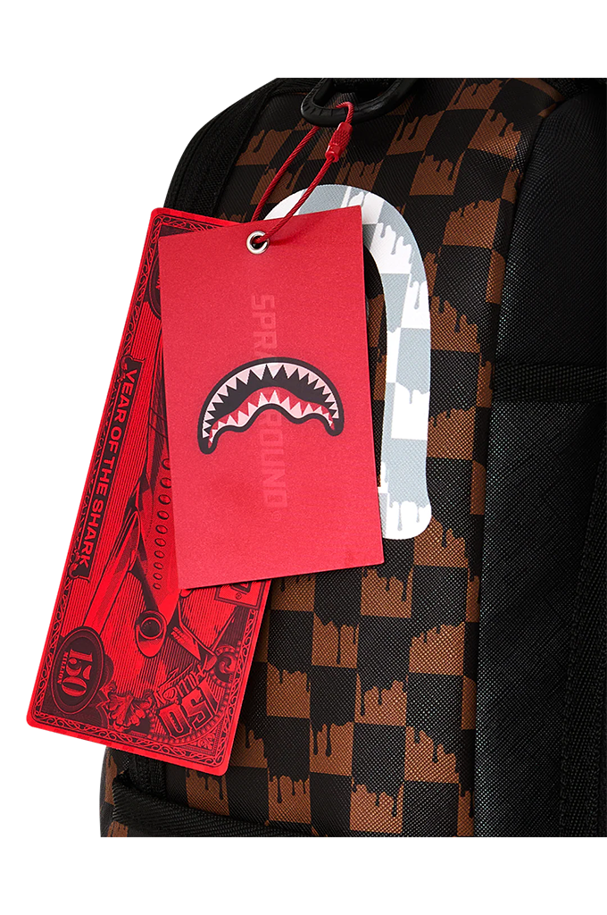 Close-up of a patterned backpack with a red hangtag featuring a shark mouth logo and the word 'SPRAYGROUND', alongside a white carabiner clip on a white background.
