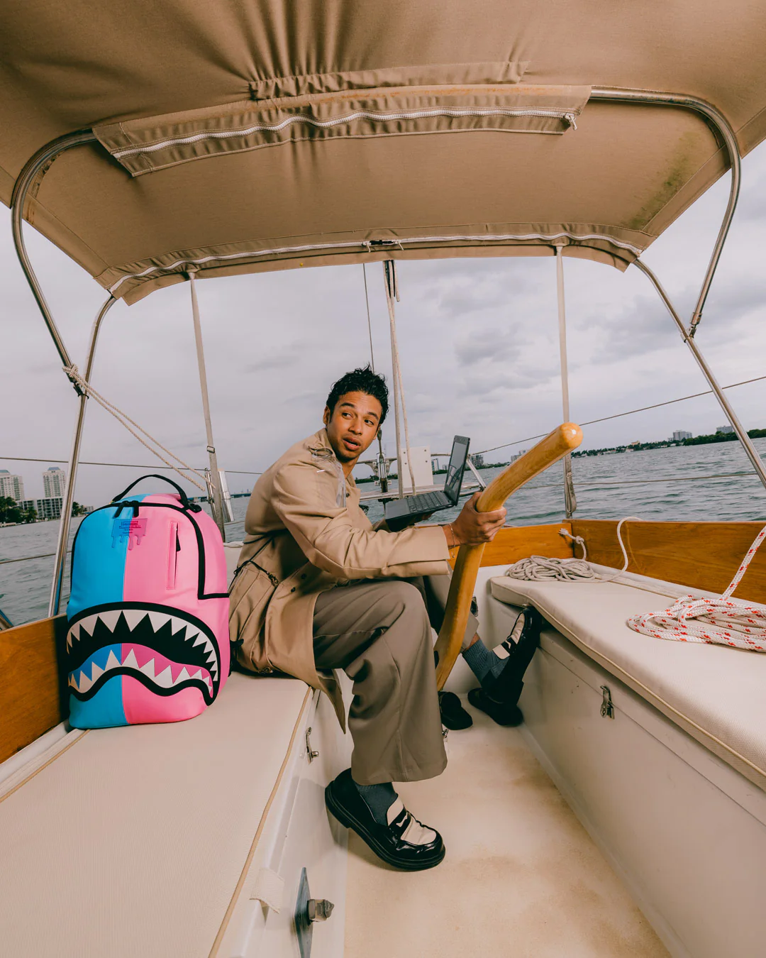 A person sitting on a boat cabin bench holding a wooden oar, with a bright pink and blue shark-mouth backpack beside them. The scene is outdoors on the water under a beige canopy.