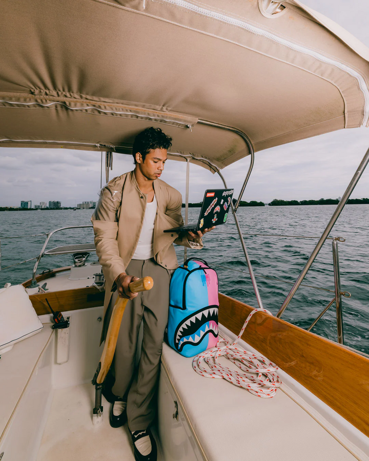 A man stands on the helm of a boat wearing a beige blazer, white t-shirt, and grey trousers, holding a laptop. A bright blue and pink shark-mouth backpack sits next to him on the boat deck, with water and a distant city skyline in the background.