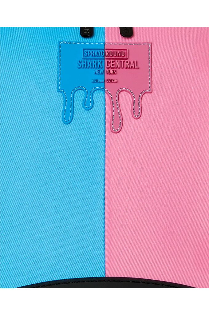Close-up of a backpack or bag panel split vertically into bright blue on the left and pink on the right, with a stitched label reading 'SPRAYGROUND SHARK CENTRAL' and a black zipper edge at the bottom.