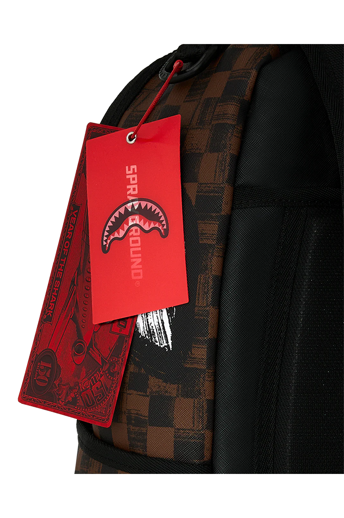Close-up of a backpack corner with a red hanging tag featuring the Shark Attack logo and the word SPRAYGROUND. The backpack has a black and brown checkerboard pattern on the side panel and black mesh fabric.