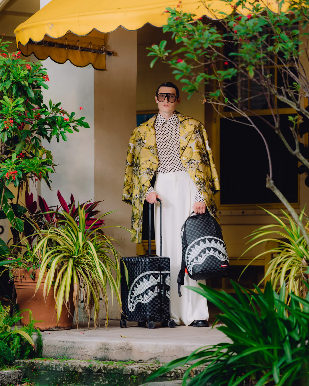 A man wearing a yellow patterned jacket, white trousers, and sunglasses stands on a porch holding a black rolling suitcase and a black backpack with a white shark graphic. The setting is lush with green plants and a yellow awning.