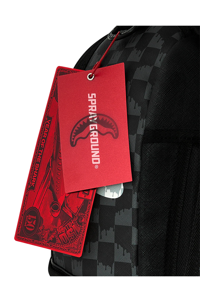 Close-up of a black and gray checkered backpack with a red paper hangtag printed with the brand name 'SPRAYGROUND' and shark graphics.