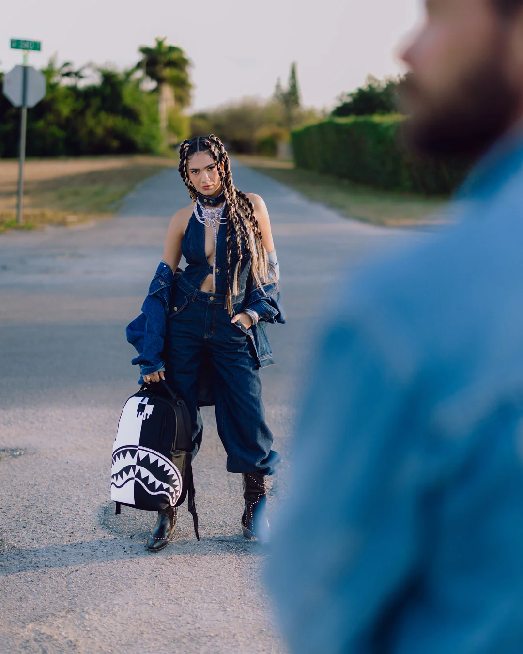 A woman stands on a paved road wearing oversized dark denim clothing and holding a black-and-white shark-mouth patterned bag. A blurred person in a blue shirt occupies the right foreground, and trees and hedges line the road behind her.
