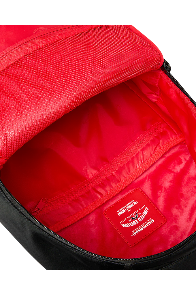 Close-up of the interior of a red backpack showing a zippered compartment, mesh lining, and a red brand patch that reads 'SPRAYGROUND'.