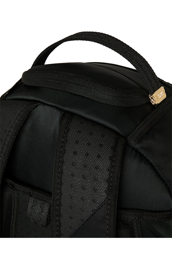 Close-up of a black backpack showing the top handle, padded zipper compartment, textured shoulder strap, and a small gold-toned metal accent on a white background.