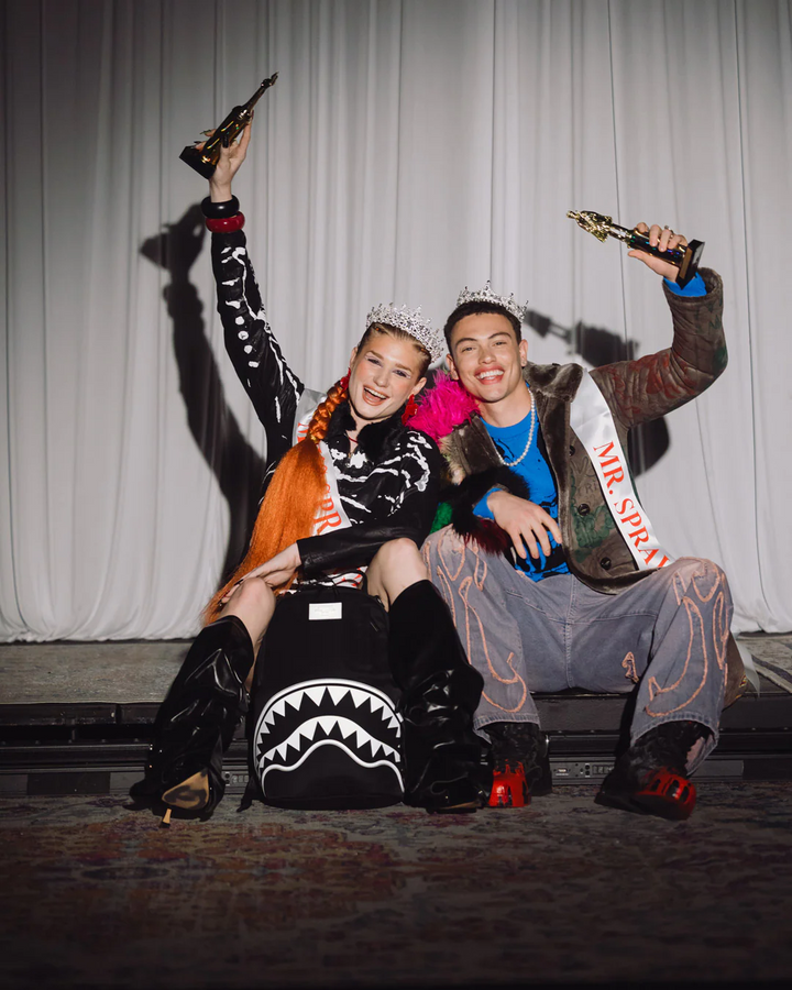 Two models posing together on a stage with awards, wearing elaborate streetwear outfits. The model on the left wears a black and white skeleton print top, a black skirt with a white shark-mouth graphic, and black boots, holding a trophy.