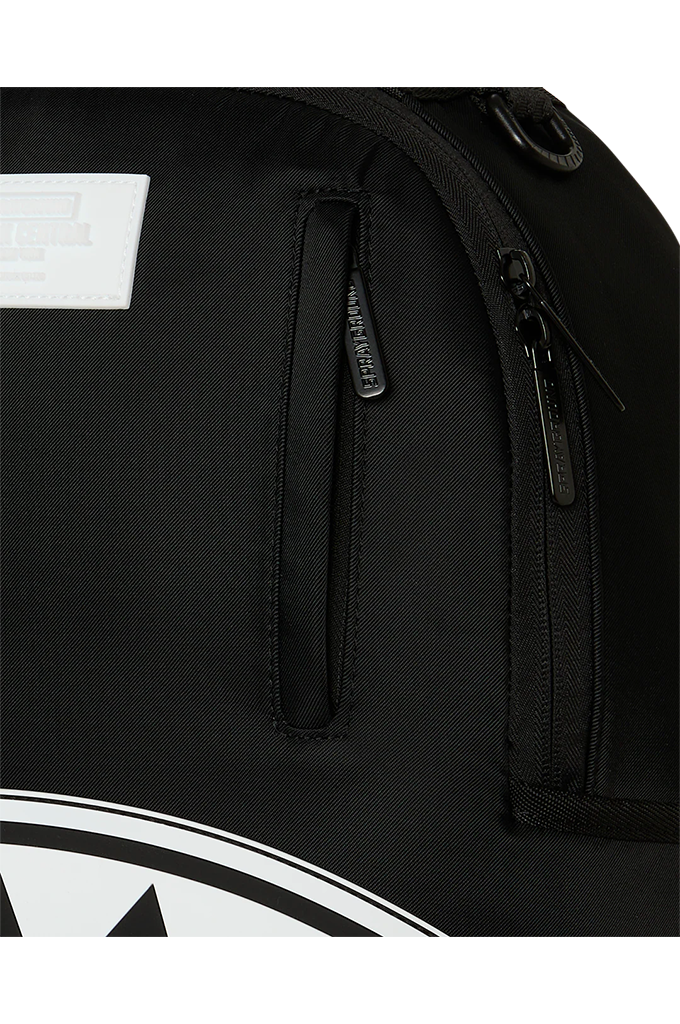 Close-up of a black backpack showing a vertical zippered pocket with a black zipper pull, a white rectangular patch on the upper left, and part of a white graphic logo at the bottom.