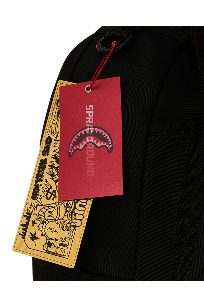Close-up of a black backpack with a red hangtag featuring a shark mouth logo and the brand name SPRAYGROUND, next to a yellow graphic paper tag.