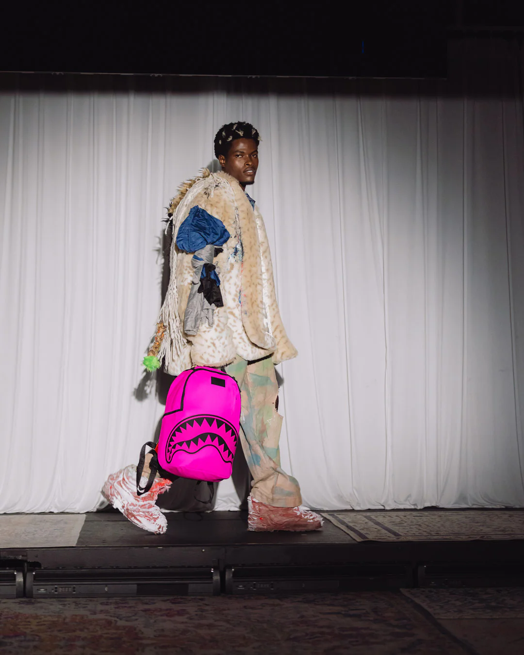A model walks a runway wearing a highly textured, patchwork-style beige and brown coat with blue accents, patterned pants, and carrying a bright pink shark-mouth backpack against a white curtain backdrop.