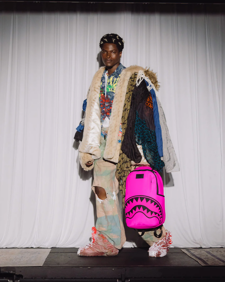 Person wearing a colorful outfit with a bright pink backpack featuring a shark design, standing against a plain background.