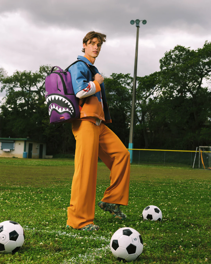 Person standing on a soccer field with a purple backpack, surrounded by soccer balls.