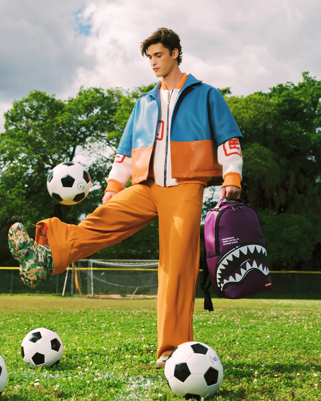 A man stands on a grassy soccer field wearing a color-blocked blue, orange, and white jacket with matching orange pants, kicking a soccer ball while holding a purple shark-mouth backpack.