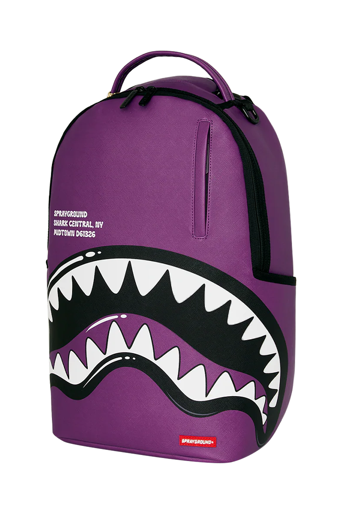 A purple Sprayground backpack featuring a large shark mouth graphic with white and black teeth across the lower front, plus a top handle and zippered compartments, shown on a plain white background.