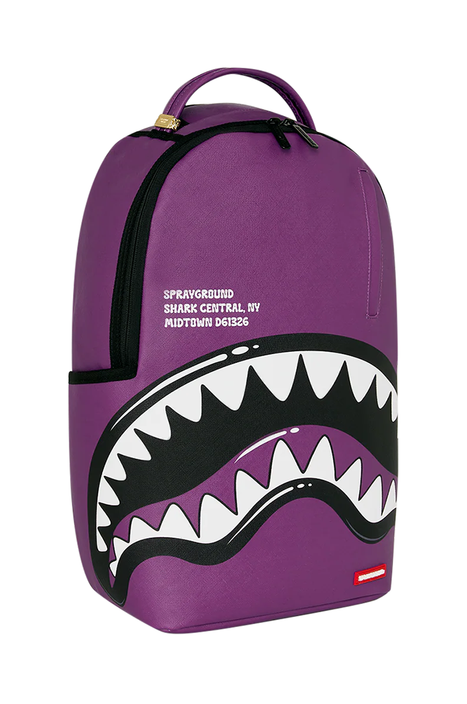A purple Sprayground Shark Central backpack featuring a large shark mouth graphic with white teeth and black outlines on the front, with Sprayground branding printed on the side.