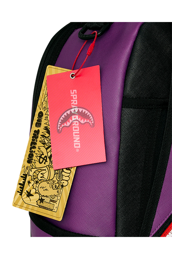 Close-up of a purple and black backpack with a red and pink 'SPRAYGROUND' shark tag and a gold tag hanging from a red string.